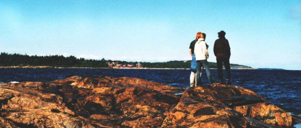 Foto: Drei junge Menschen stehen auf einem flachen Felsen in Bjuroklübb an der Ostküste Schwedens, drehen dem Betrachter den Rücken zu und schauen aufs Meer hinaus. Im Hintergrund sieht man auf einer Landzunge ein kleines Dorf umgeben von Tannenwald.